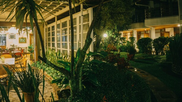 Un espacio exterior bien iluminado con zona de comedor, rodeado de abundante vegetación y un sendero que lleva a un edificio con grandes ventanales.