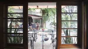 Vista a través de una ventana de madera con múltiples divisiones, que muestra un área de estar exterior con mesas y sillas, bicicletas estacionadas al frente y algunas personas sentadas y de pie.