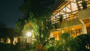 Un edificio moderno con fachada de madera y ventanas amplias, rodeado de exuberante vegetación y un espacio exterior bien iluminado.