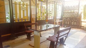 Un espace repas avec une table et des bancs en bois, une enseigne indiquant « SWEET », et une vue à travers une clôture à treillis.