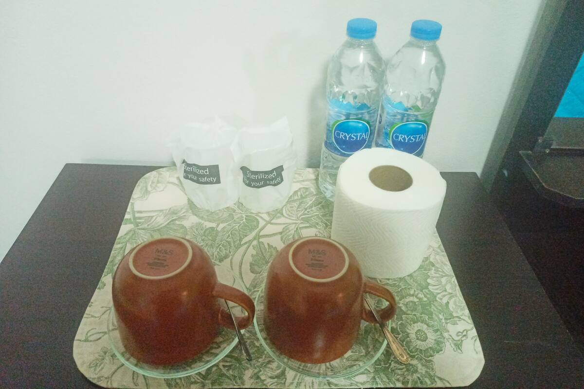Un plateau contenant deux tasses brunes, deux bouteilles d’eau, deux sachets stérilisés et un rouleau de papier toilette, le tout posé sur une nappe à motifs.
