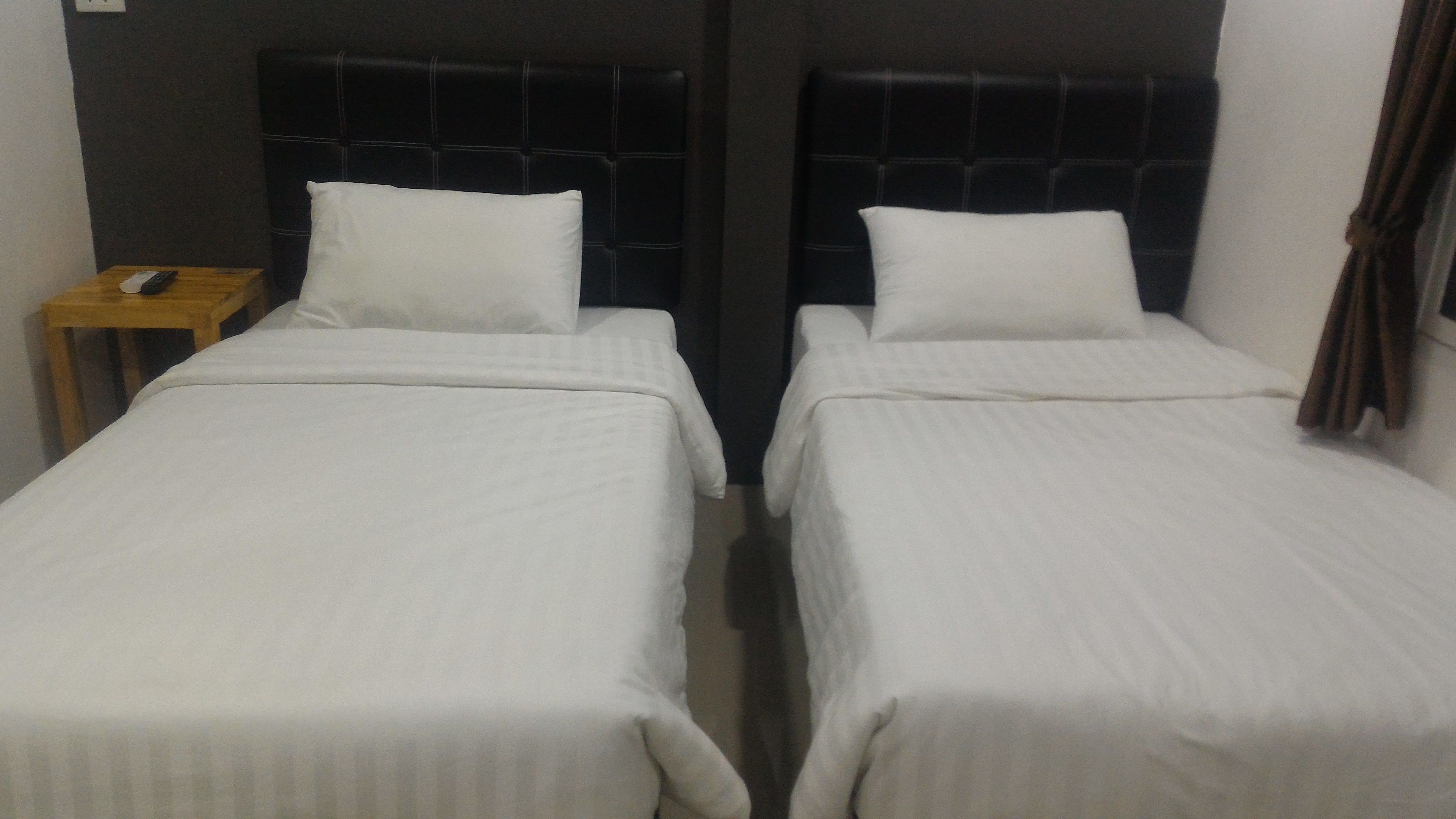 Ein Zimmer mit zwei Einzelbetten, jeweils mit weißer Bettdecke und Kopfkissen, einem hölzernen Nachttisch mit Fernbedienung und einem dunklen Kopfteil.