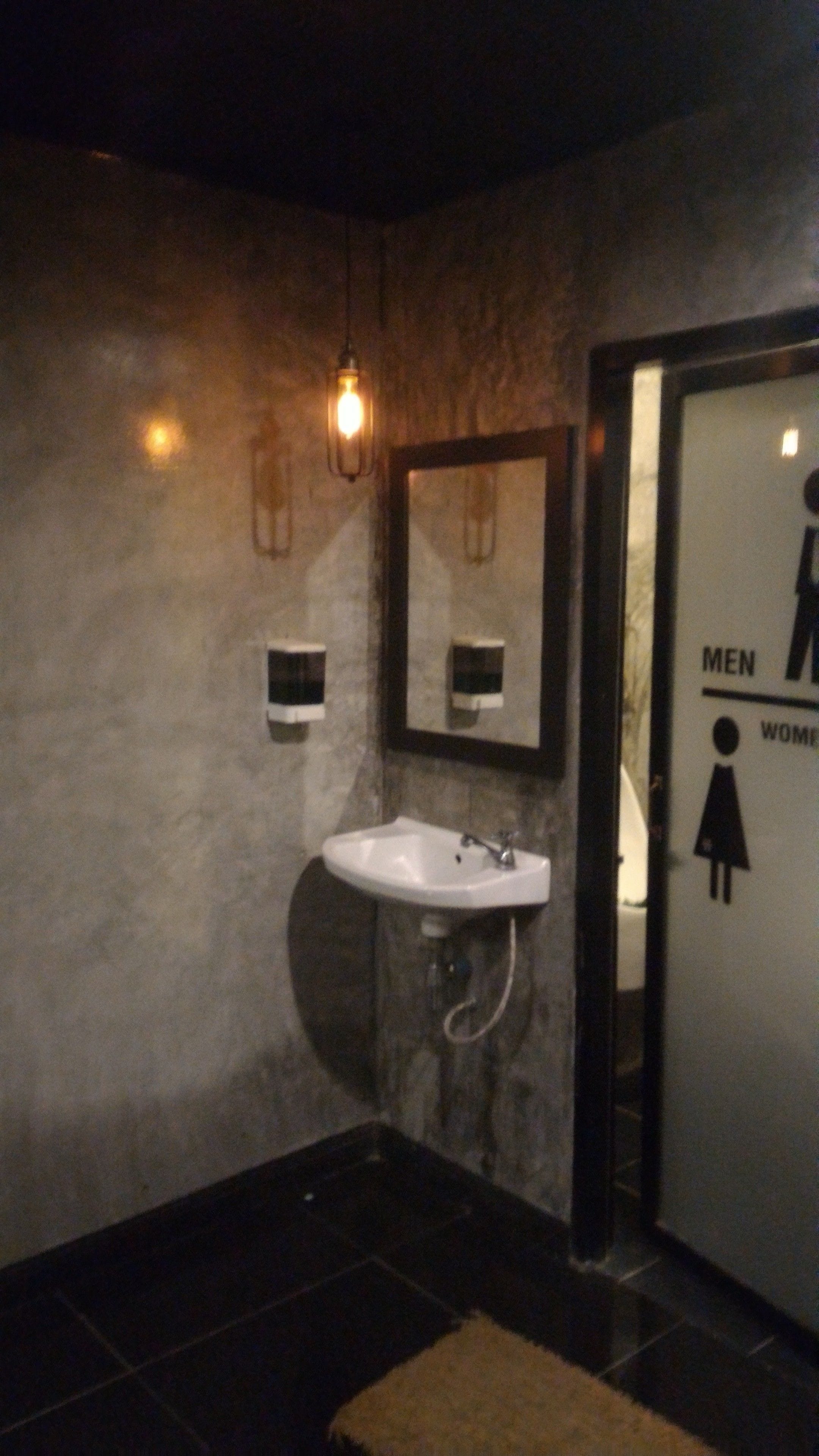 Une salle de toilette avec un lavabo blanc, un miroir et un luminaire fixé au mur. Des panneaux indiquent les toilettes séparées pour hommes et pour femmes.
