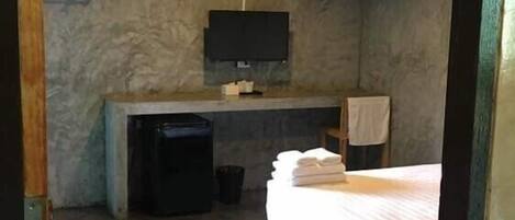 Habitación de hotel compacta con una cama, un escritorio pequeño, un televisor montado en la pared y un cesto de basura.