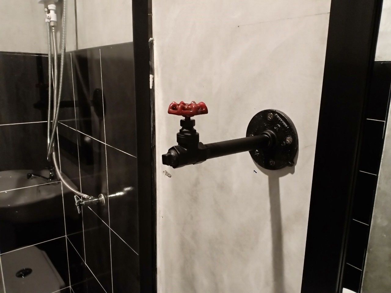 Baño con pared de azulejos blanco y negro, cabezal de ducha manual y grifo montado en la pared con pomo rojo.
