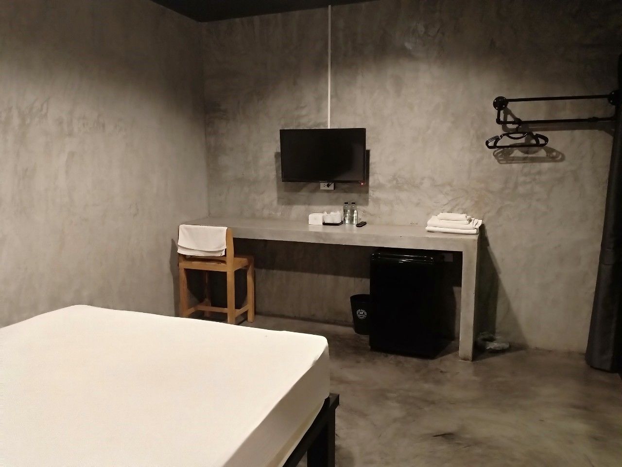 Una habitación pequeña y tenuemente iluminada con una cama, un escritorio, una silla, un televisor y un basurero.