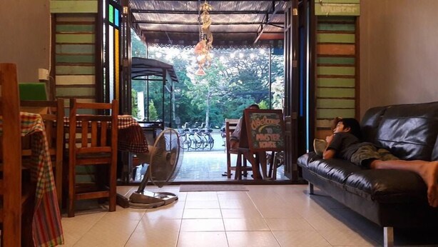 An indoor seating area with a view of bicycles outside, a fan on the floor, and a signboard. - Muster House - Hostel (Ayutthaya)