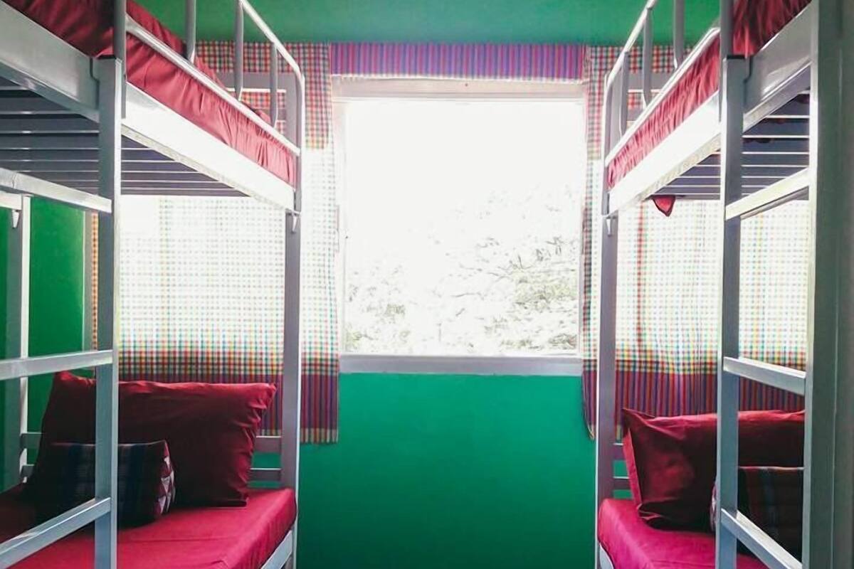 A bunk room with two bunk beds, each with red bedding and pillows, a window with a checkered curtain, and an air conditioning unit above.