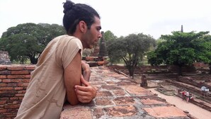 A person leaning on a stone wall at an archaeological site with ancient ruins and trees in the background. - Muster House - Hostel (Ayutthaya)