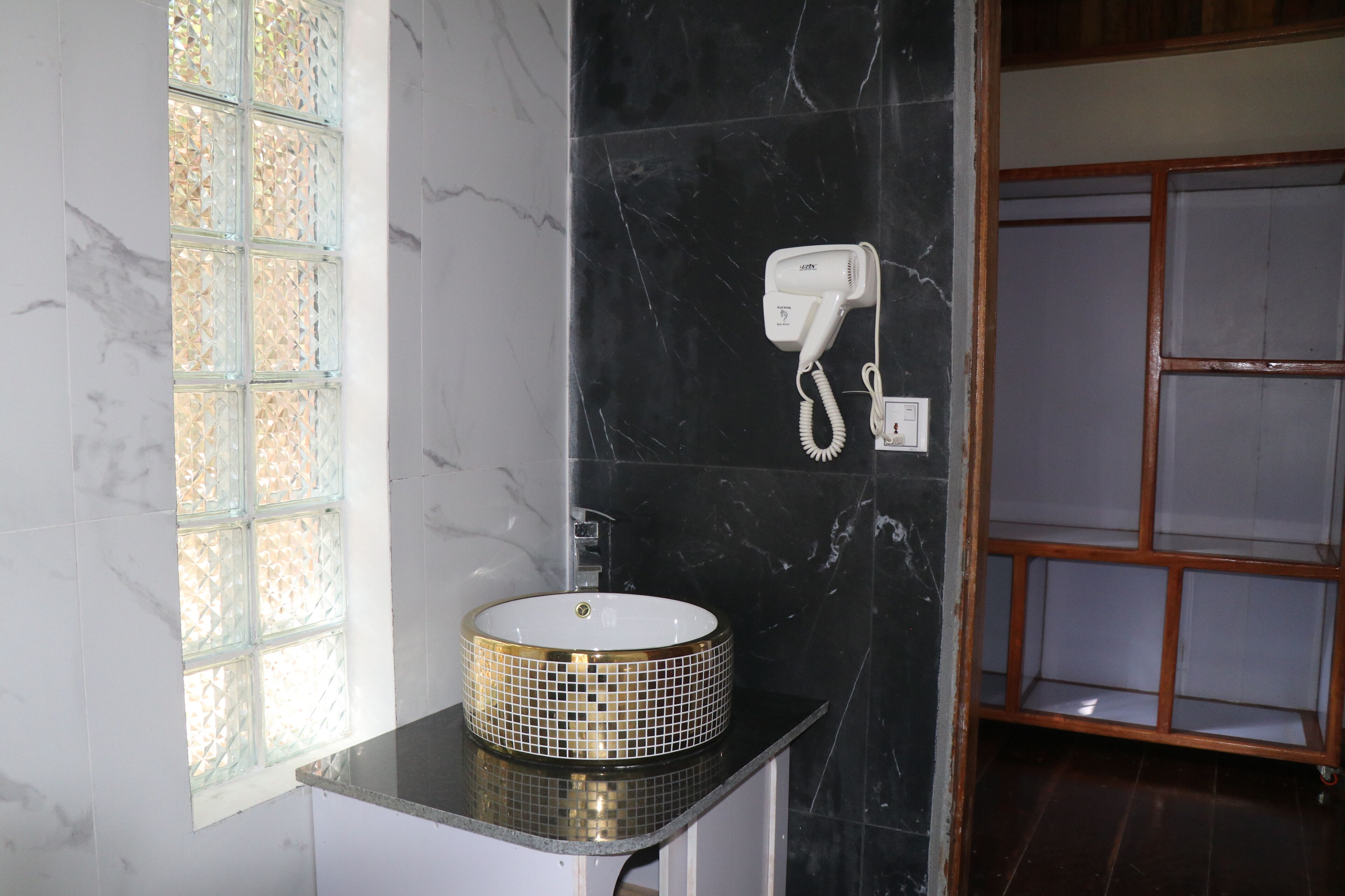 A bathroom with a marble wall, a glass window, a sink with a gold rim, and a hairdryer mounted on the wall.