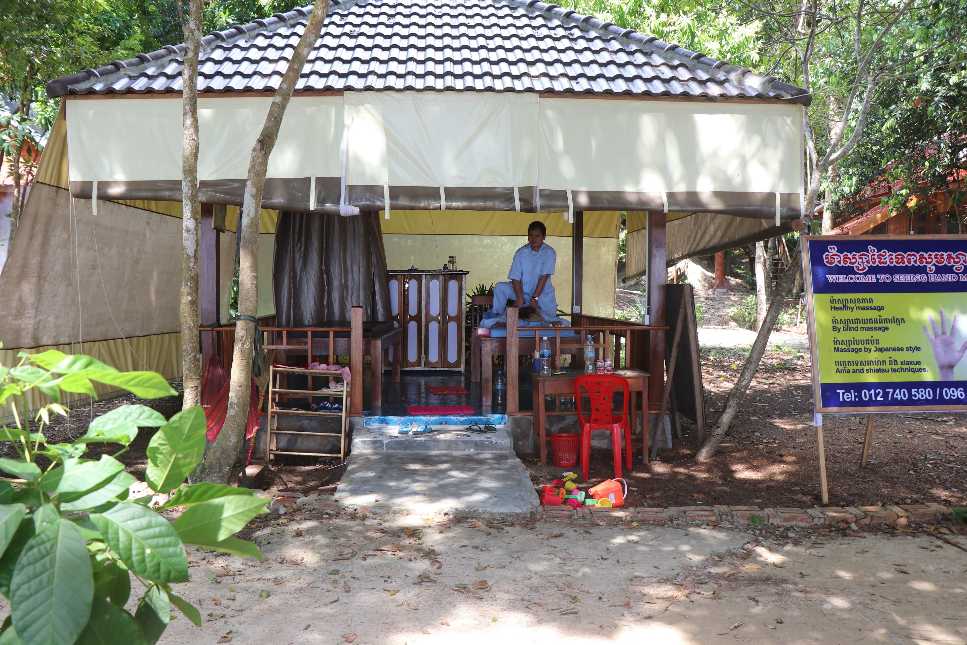 A small, single-story structure with a tiled roof and a covered seating area. A person is seated at a table under a canopy.