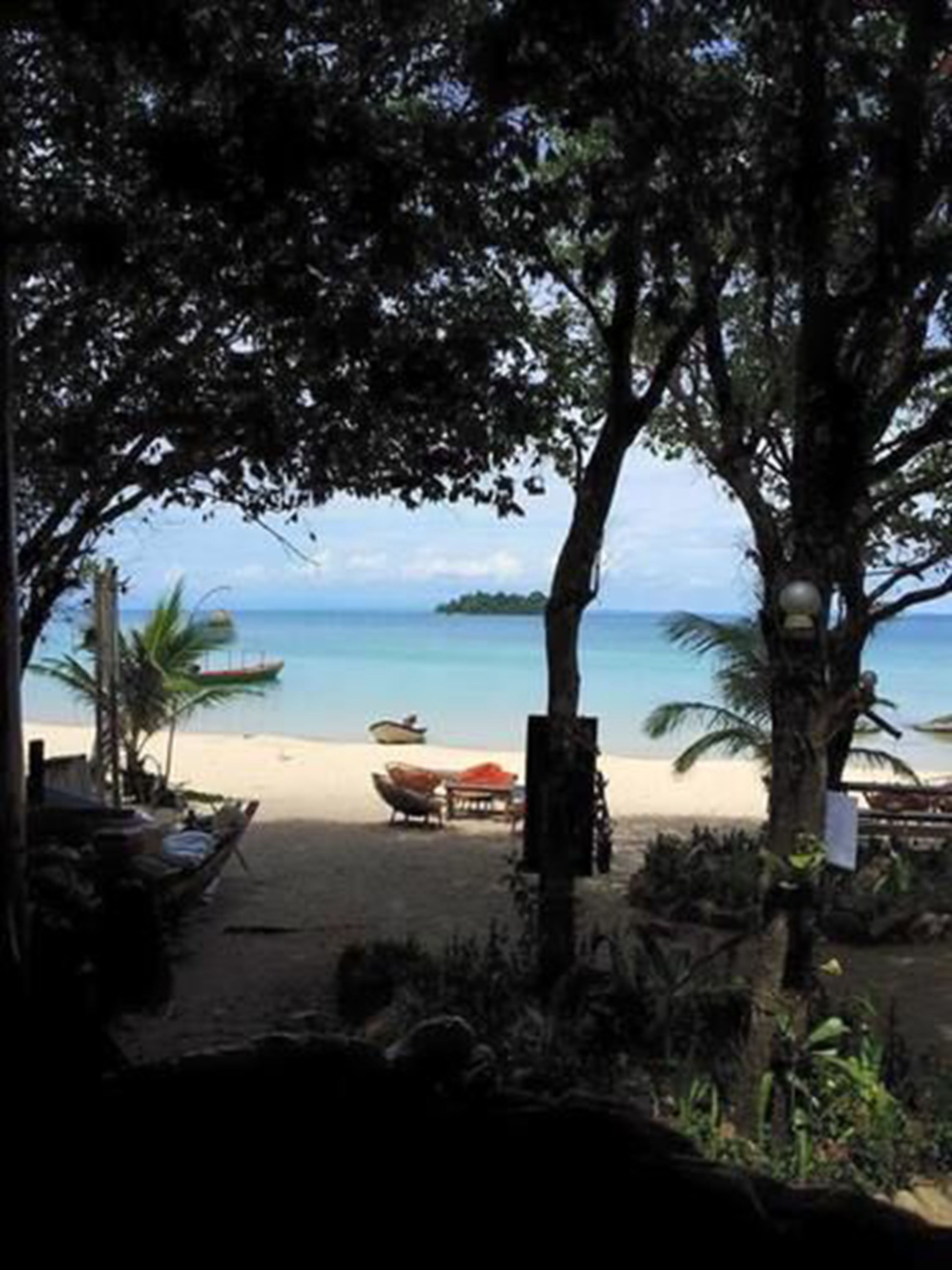 Vista a la playa con un bote, una mesa y sillas bajo un árbol.