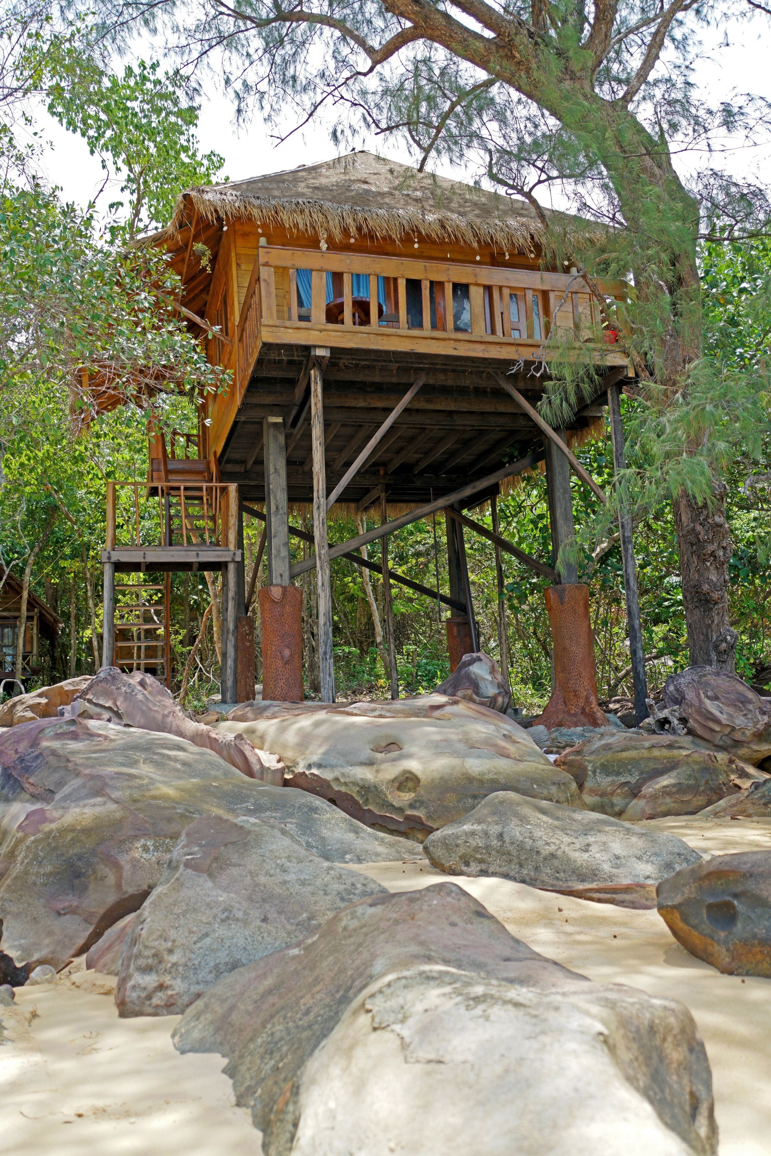 Una casa en árbol de madera con techo de paja, construida sobre pilotes, rodeada de rocas grandes y árboles.