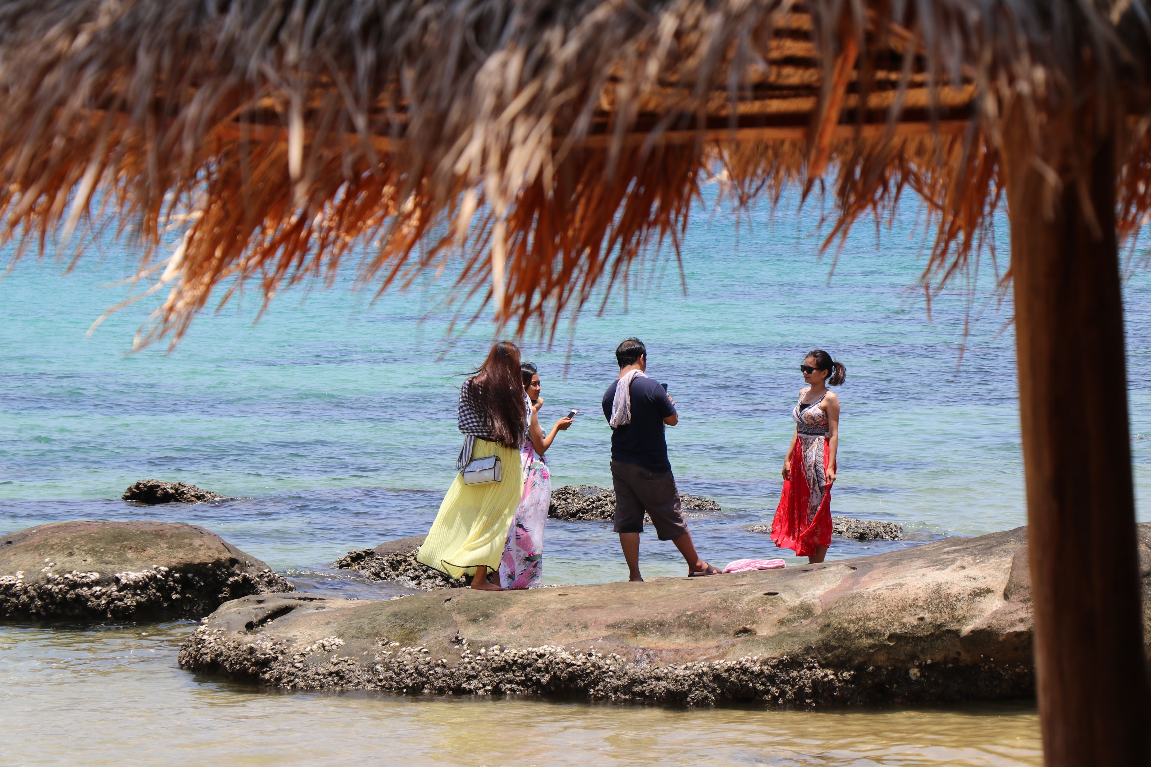 People standing on rocks by a body of water, with a thatched structure overhead.