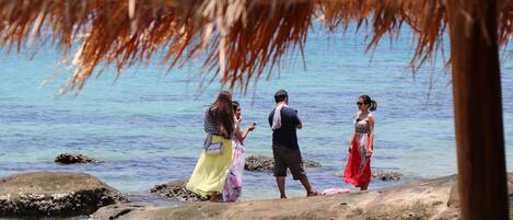 People standing on rocks by a body of water, with a thatched structure overhead.