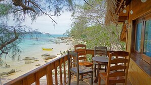 A wooden deck with a table and chairs overlooking a beach with clear water and rocks. - Tree House Bungalows Resort (Koh Rong)