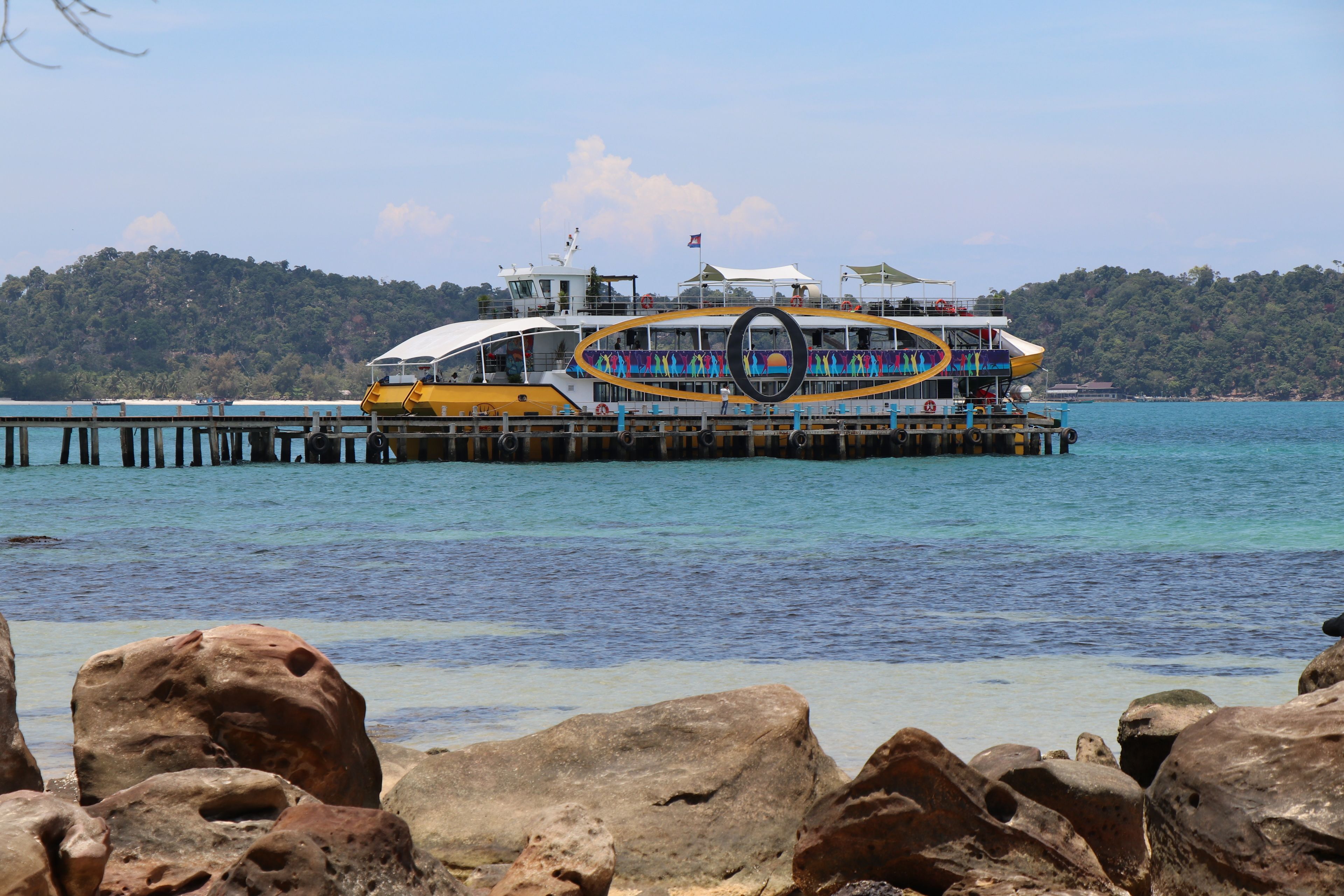 A ferry with a colorful deck and a large circular structure in the center, docked at a pier with rocky foreground and a forested hill in the background.