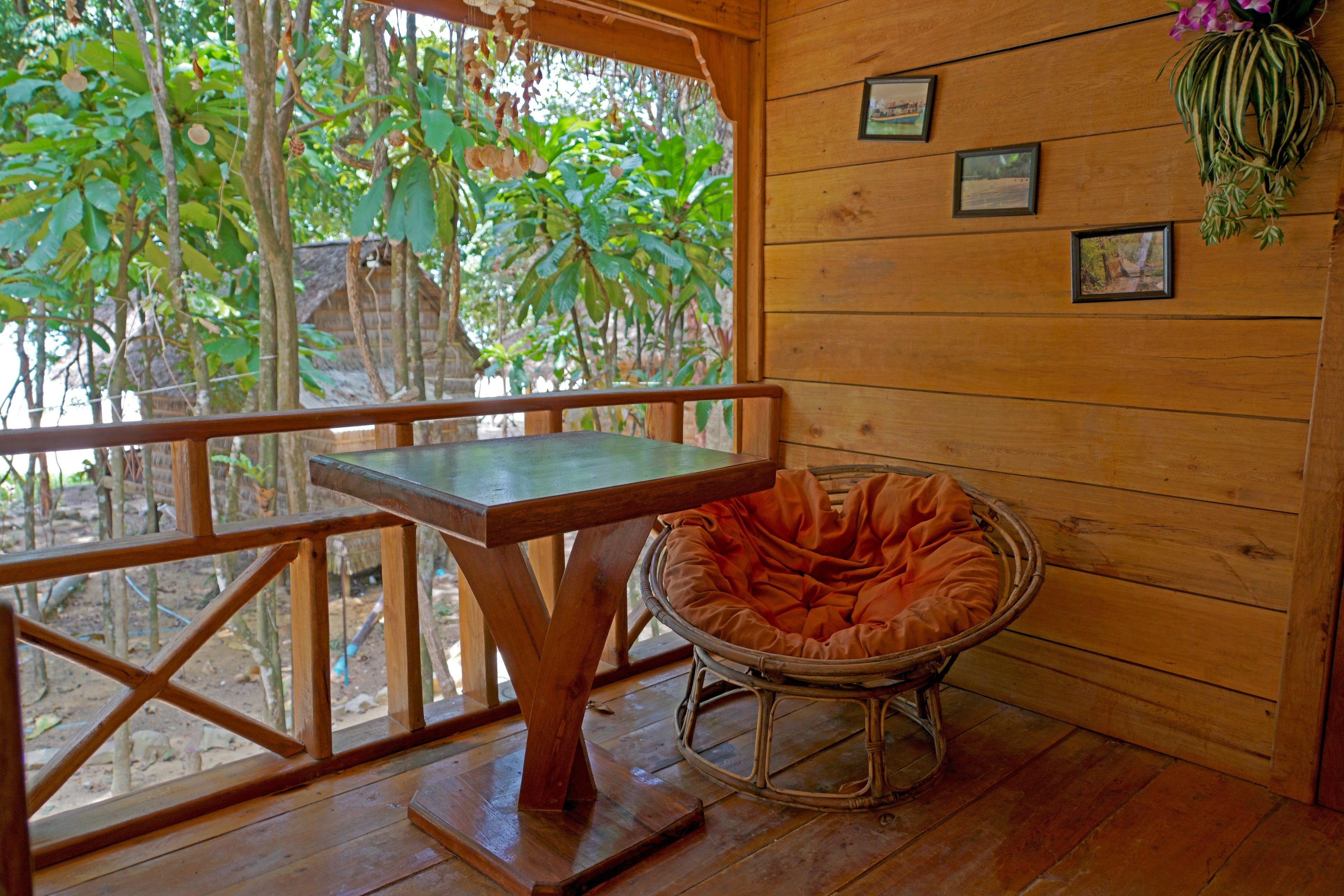 A wooden room with a table and a wicker chair, a wooden railing, and a window overlooking greenery.