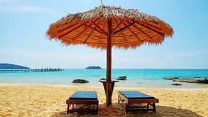 A beach with two wooden lounge chairs under a thatched umbrella, a wooden pier in the background, and clear blue water. - Tree House Bungalows Resort (Koh Rong)