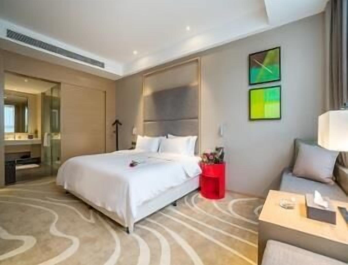 A modern hotel room with a large bed, a seating area, a red side table, and a large mirror.
