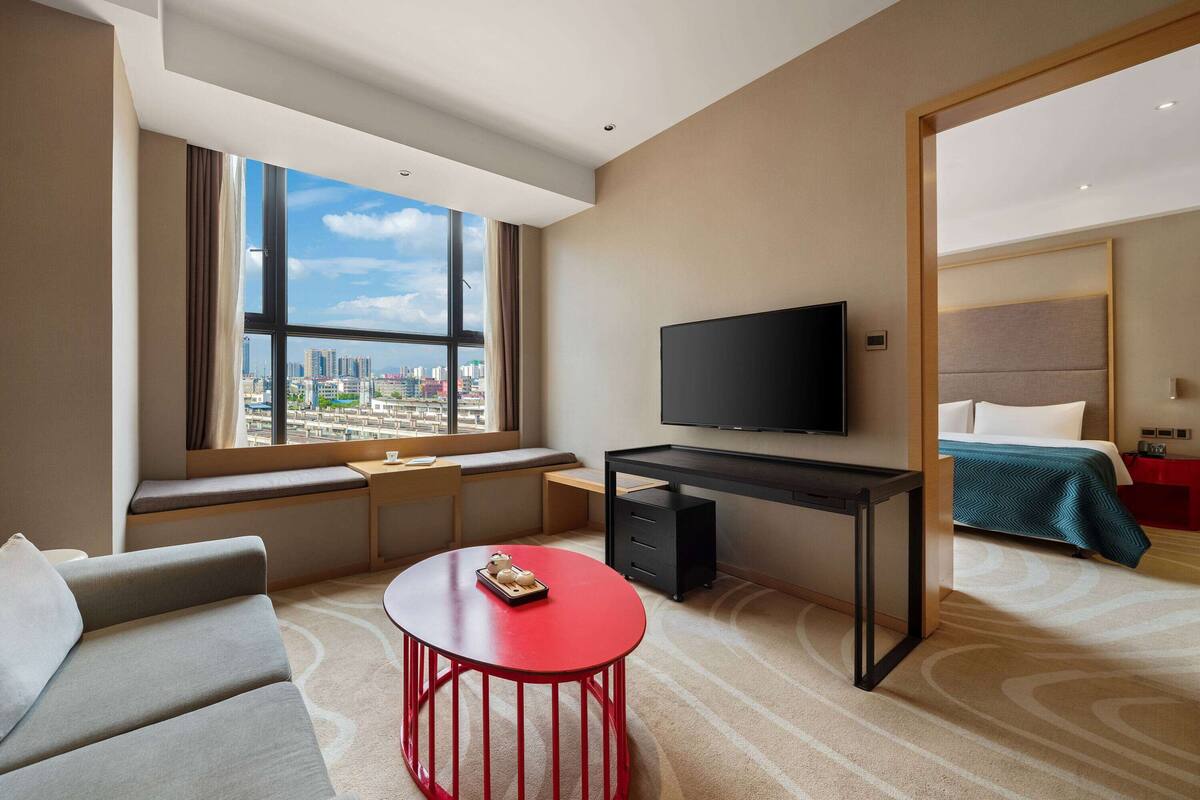 Una habitación de hotel moderna con un ventanal grande, un televisor de pantalla plana, un escritorio, una mesa de centro roja, un sofá y una cama con colcha azul.