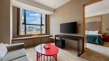 Una habitación de hotel moderna con un ventanal grande, un televisor de pantalla plana, un escritorio, una mesa de centro roja, un sofá y una cama con colcha azul.