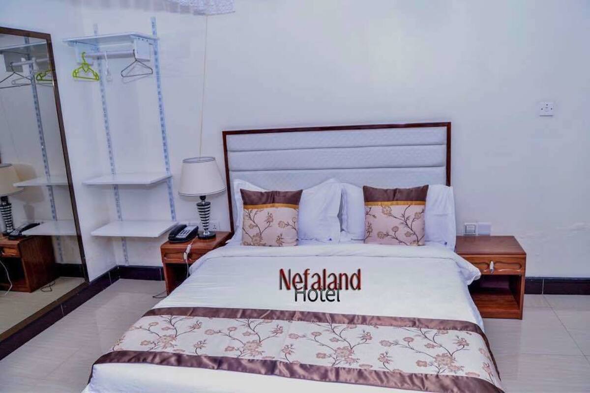 Een net opgemaakt bed met een bloemenprint beddeken, een bedlampje en een nachtkastje met een telefoon. De kamer heeft een spiegel, een plank met kledinghangers, en een bordje met de tekst 'Nefaland Hotel'.
