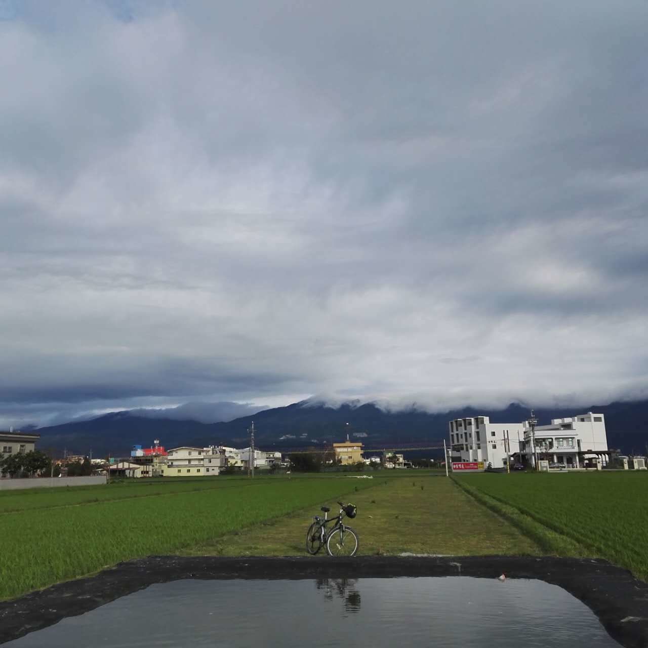 Paisaje rural con una bicicleta estacionada cerca de una masa de agua, rodeado de campos y edificios al fondo.