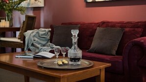 A wooden table with a tray holding a crystal decanter, two wine glasses, and a book. A red velvet sofa with a cushion is in the background. - Impressions Studio (Daylesford)