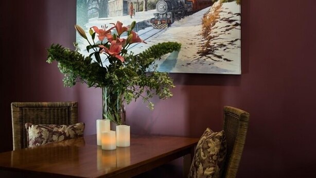 A dining table with a vase of flowers and candles, flanked by wicker chairs with patterned cushions. - Impressions Studio (Daylesford)