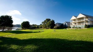 A large, two-story building with a balcony on the upper floor, situated in a grassy area with trees and a lake in the background. - Lakeside Suites 2 (Daylesford)