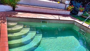 Piscina cristalina con escalones de acceso, rodeada de una terraza con baldosas y un pequeño jardín con flores.