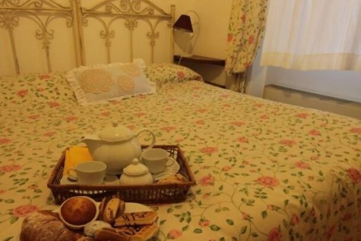 A neatly made bed with floral bedding, a tray of breakfast items including bread, pastries, and tea, and a bedside table with a lamp.