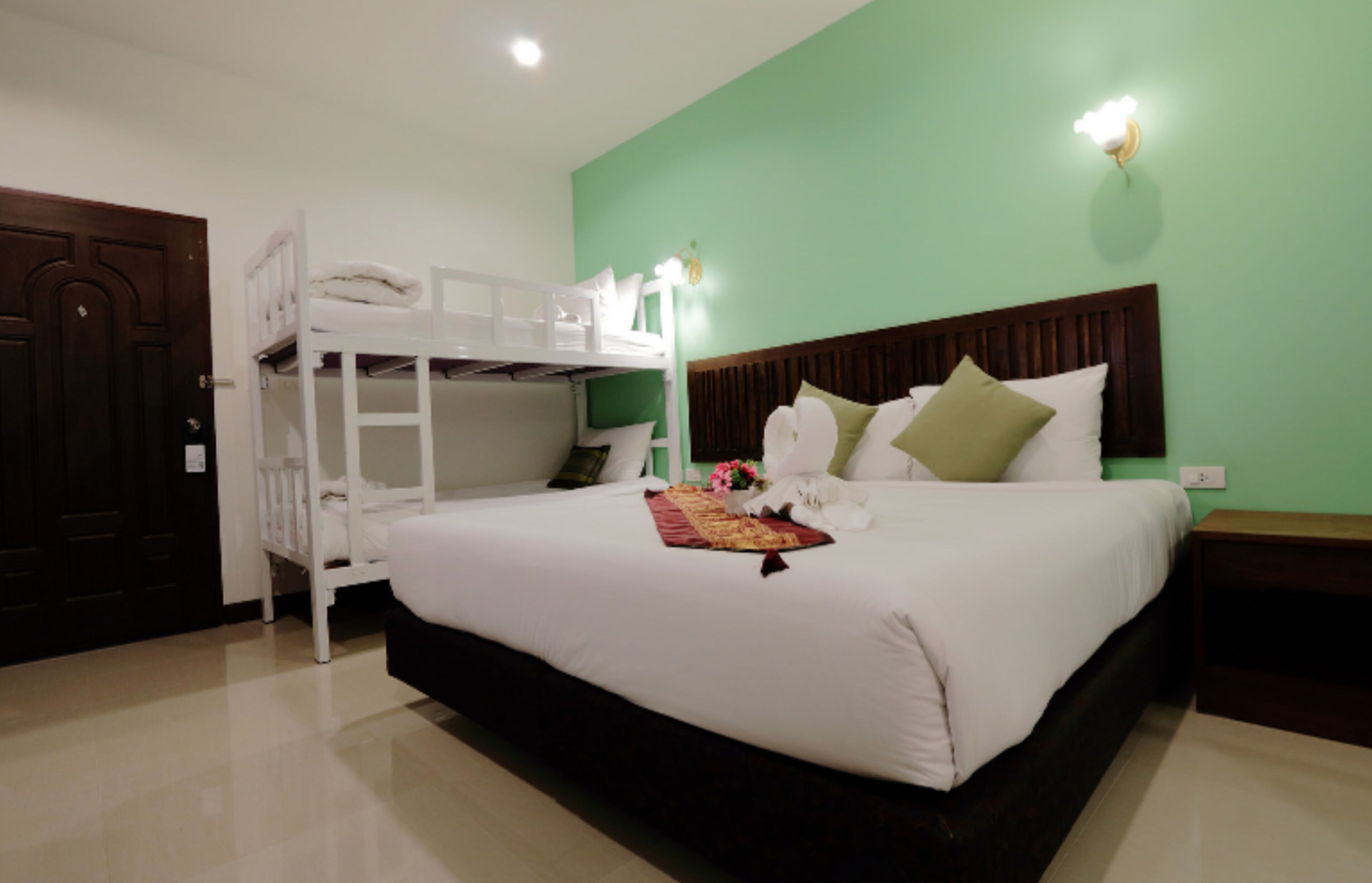 A hotel room with a bunk bed, a large bed with pillows, a bedside table, and a green wall.