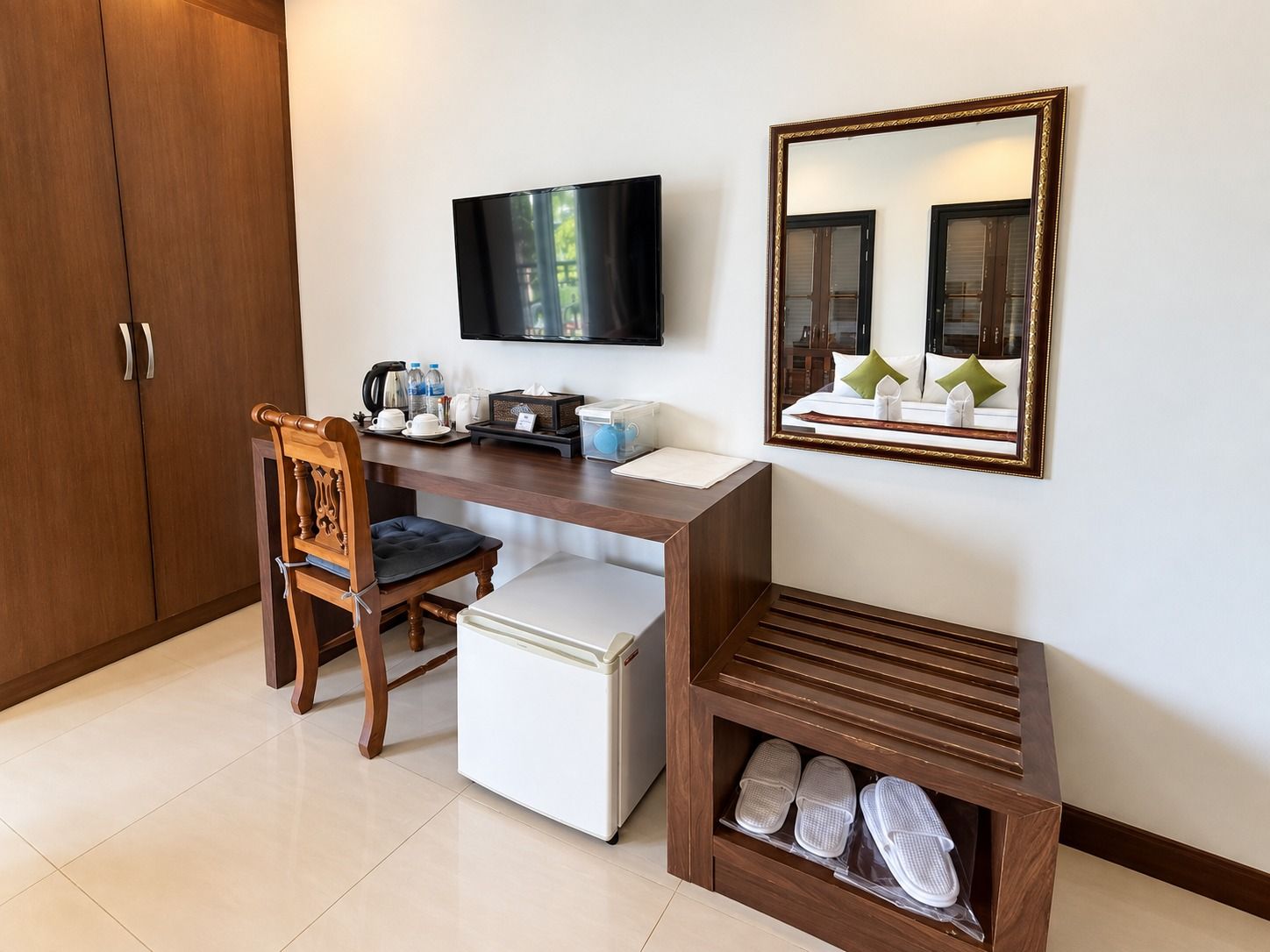 A hotel room with a wooden desk, a television, a mirror, a chair, a small white refrigerator, and a shoe rack.