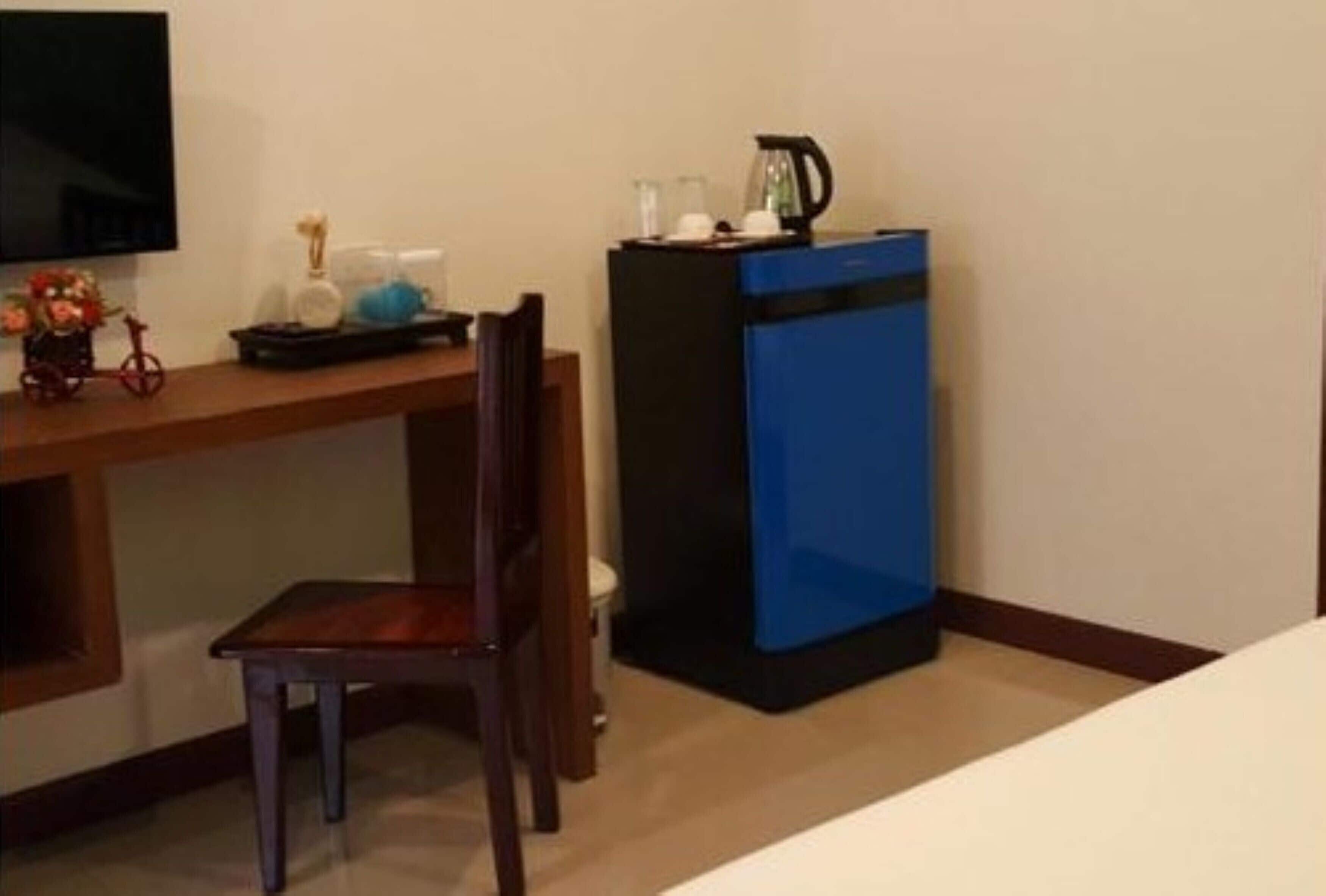 A hotel room with a wooden desk, a chair, a small table with a decorative item, a blue minibar, and a television mounted on the wall.