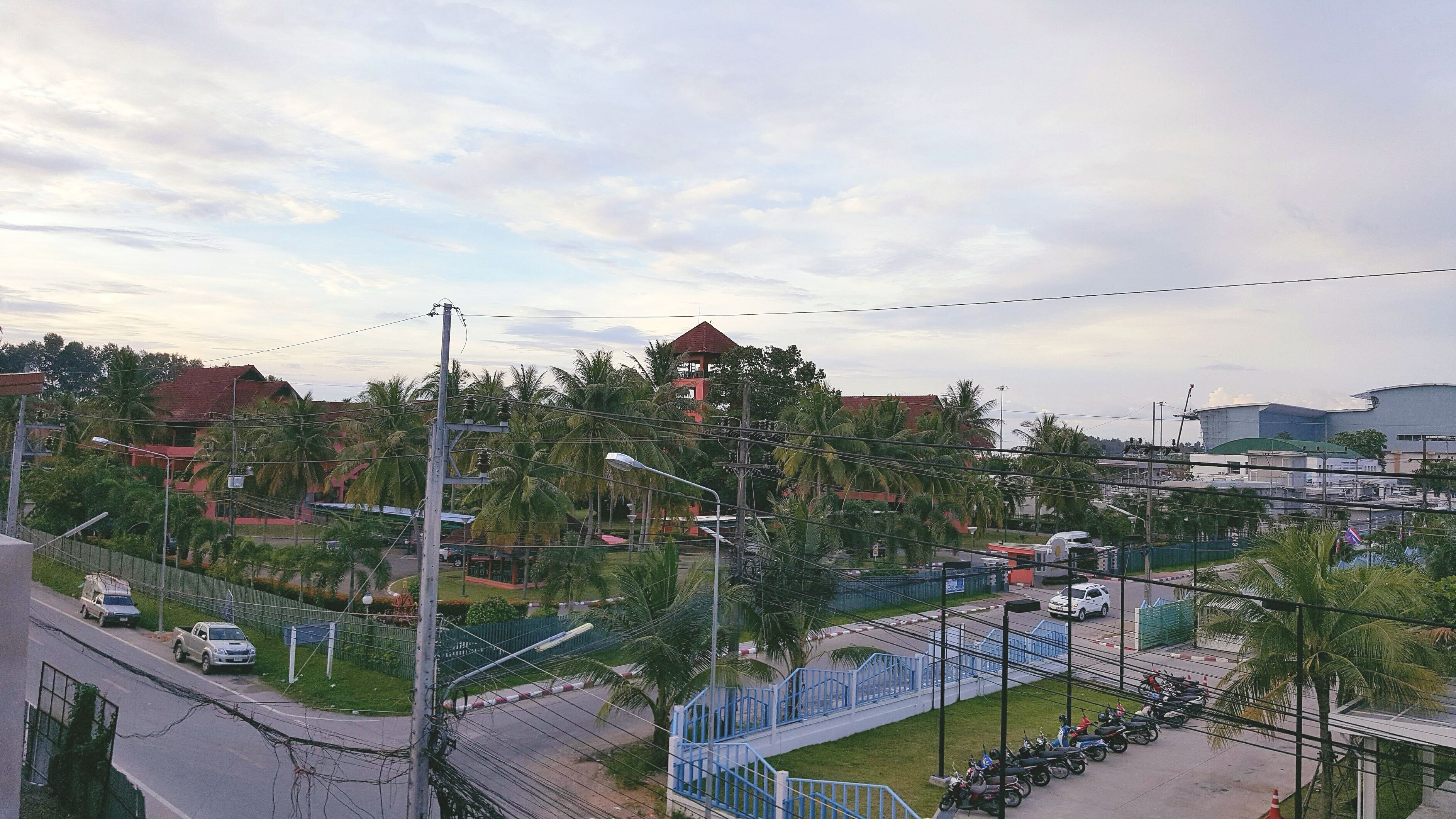 A view of a residential area with a prominent red-roofed tower, a modern building, and a street with parked vehicles and a pedestrian walkway.