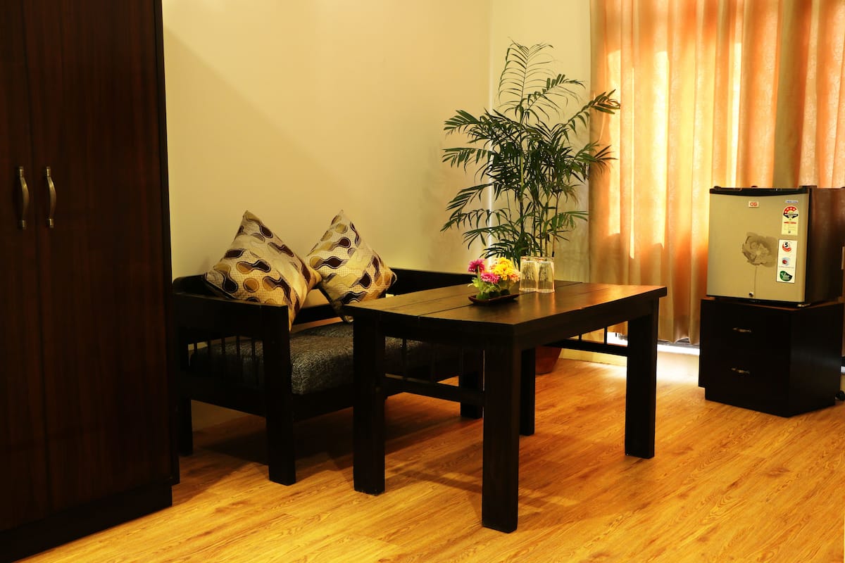A room with a wooden floor, a sofa with patterned cushions, a table with a vase of flowers, a potted plant, and a refrigerator.