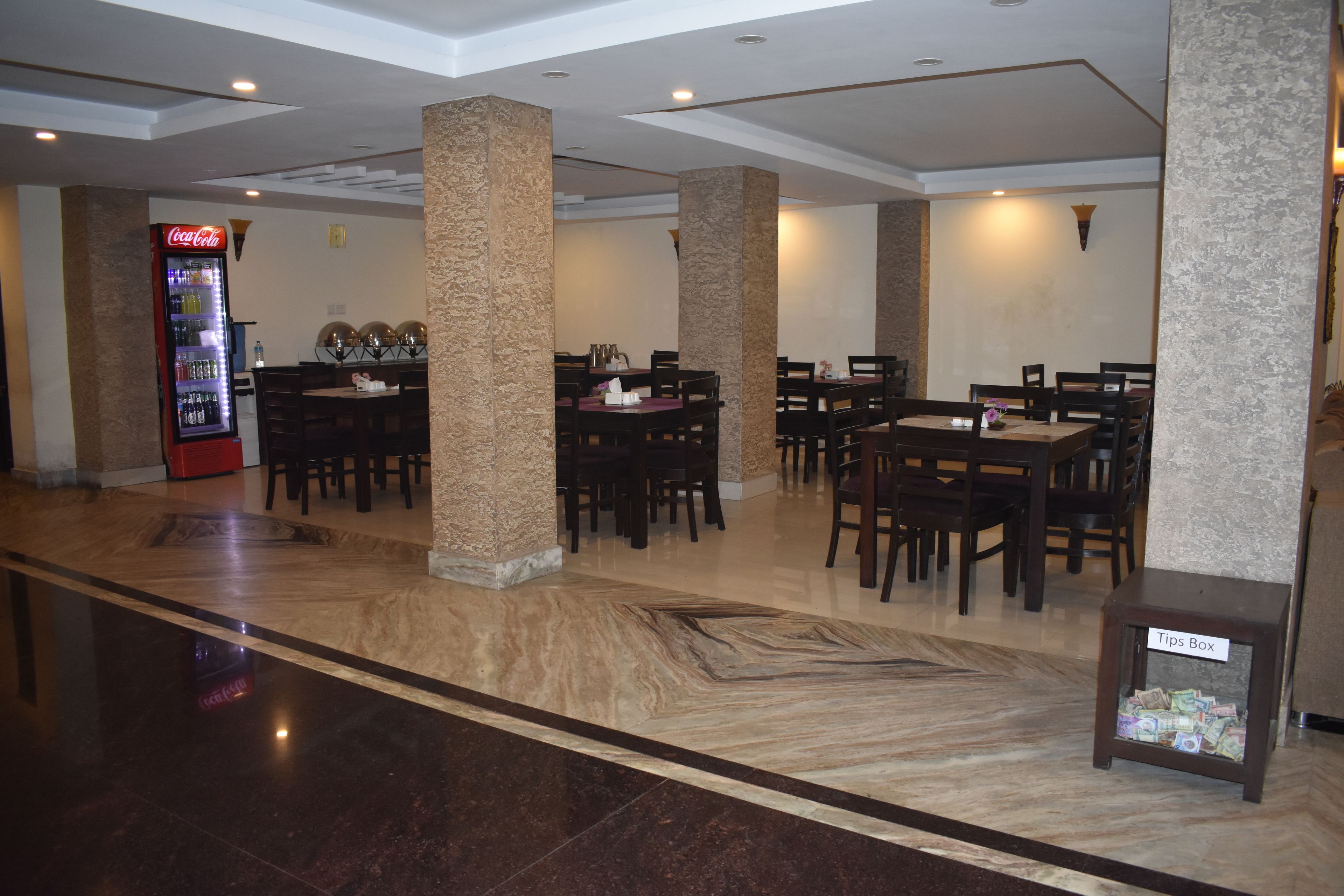 A hotel lobby with marble flooring, multiple dining tables, and a Coca-Cola refrigerator.