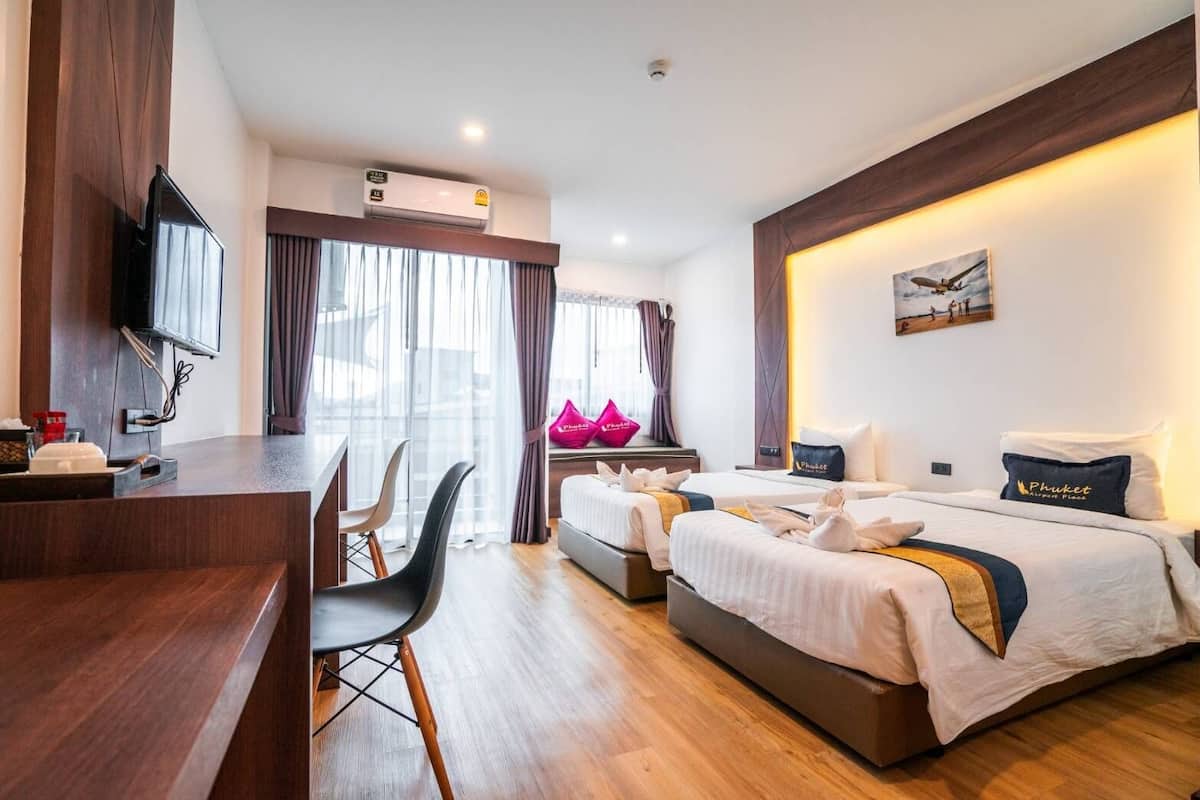 A hotel room with a wooden desk, two beds, a TV, and a large window with curtains.