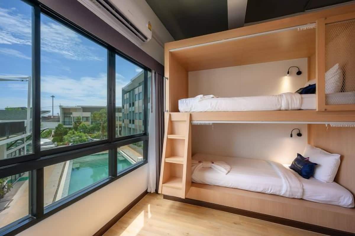 A room with two bunk beds, a large window with a view of buildings and a swimming pool, and a small shelf with a lamp.