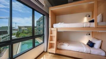 Una habitación con dos camas literas, un ventanal con vistas a edificios y una piscina, y un estante pequeño con una lámpara.