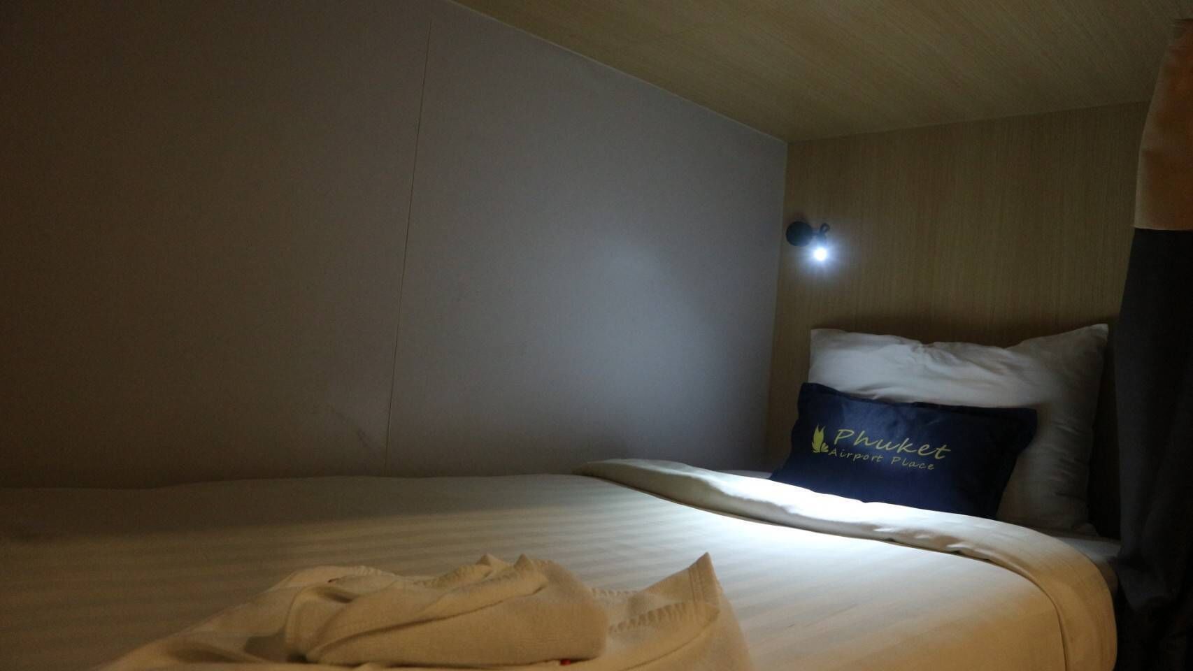 Ein Einzelbett mit einem Kissen mit dem Aufdruck 'Phuket Airport Place', einer Nachttischlampe und einem ordentlich gefalteten weißen Bademantel.