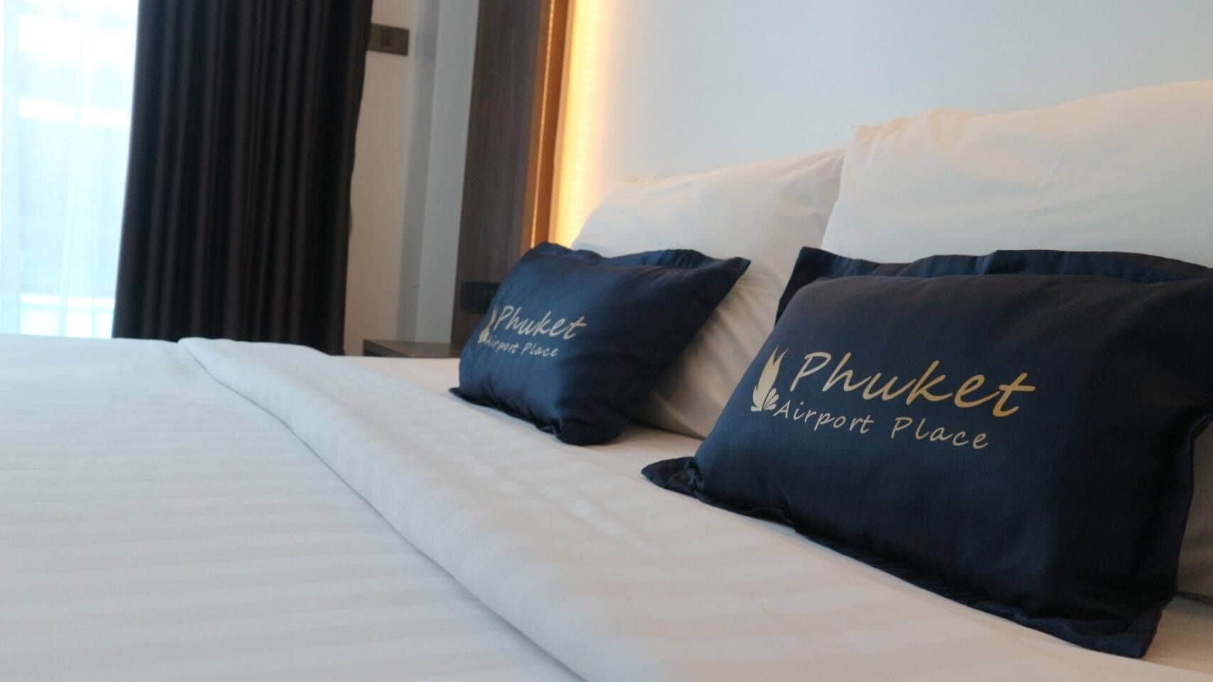 Ett hotellrum med en säng, två kuddar med texten ”Phuket Airport Place” och ett fönster med gardiner.
