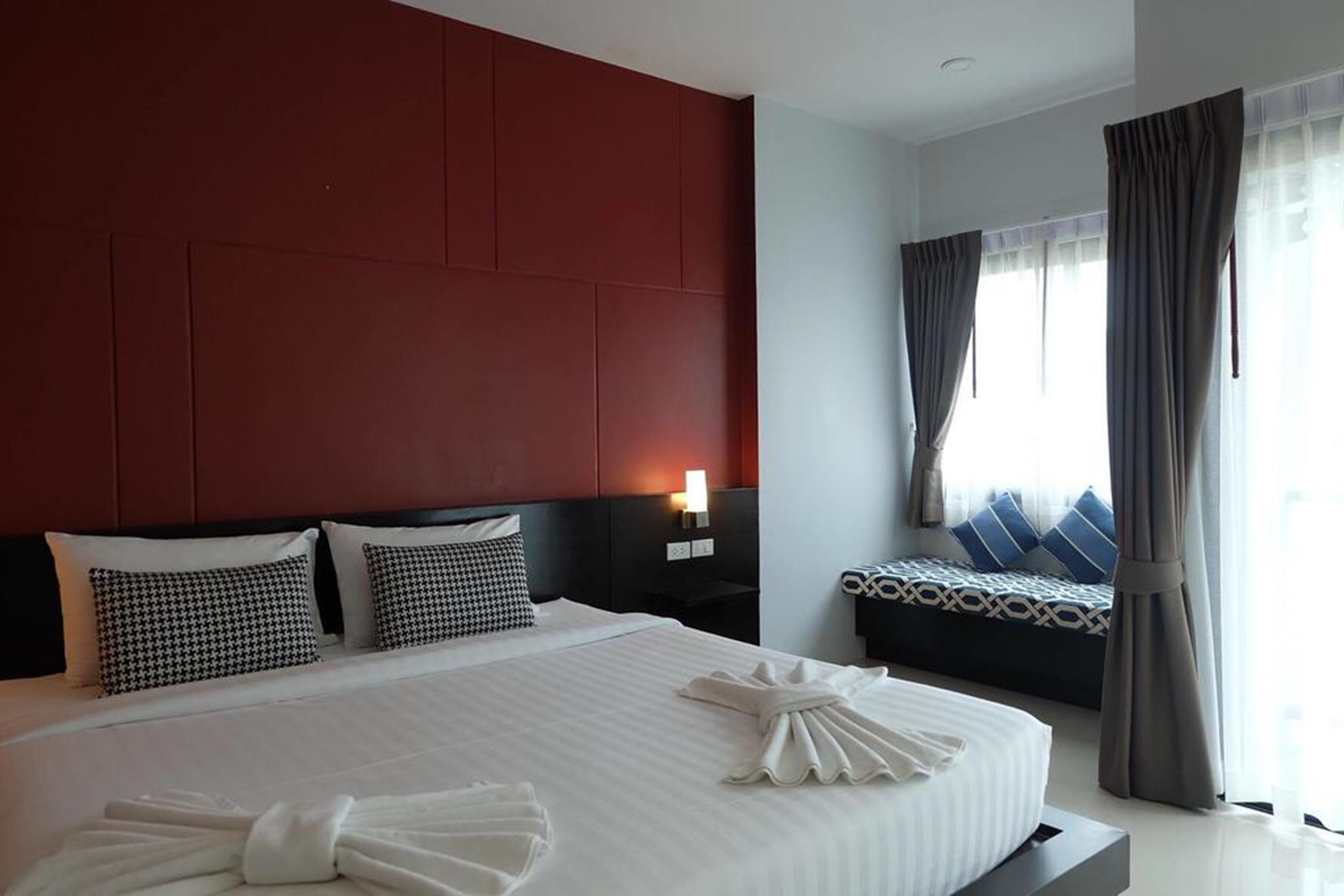 A hotel room with a large bed, a bedside table with a lamp, a smaller bed with patterned bedding, and a window with curtains.