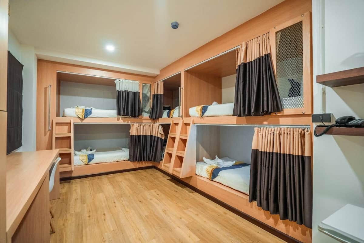 A dormitory room with bunk beds, wooden desks, and a small kitchenette area.