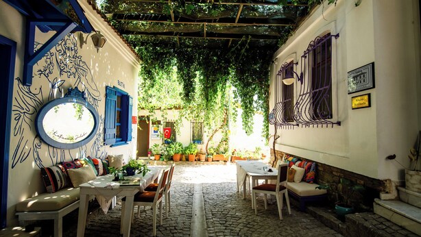 A narrow alleyway with a café setup featuring outdoor seating, a round mirror, and potted plants. - Katina Bozcaada (Bozcaada)
