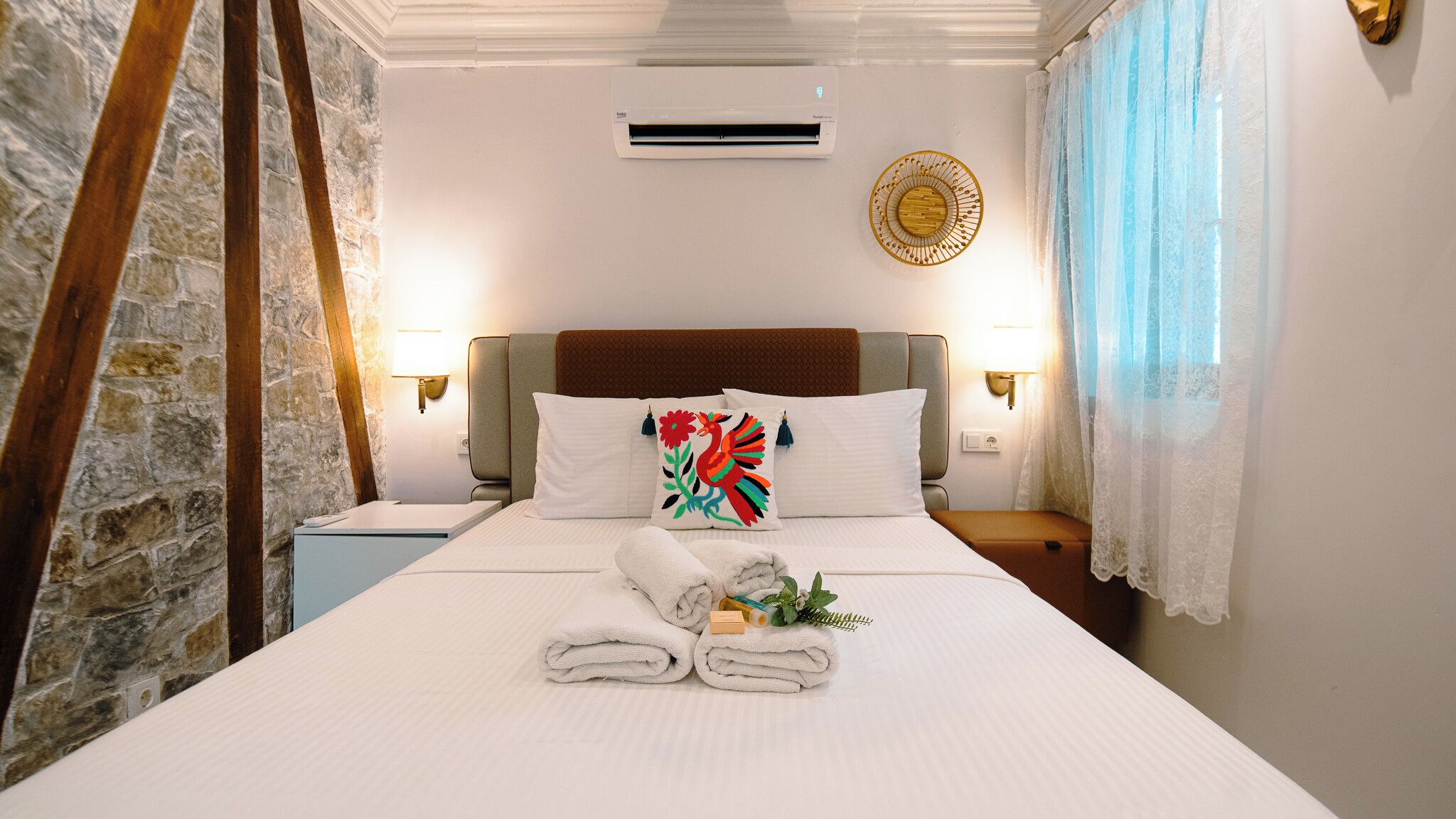 A hotel room with a bed, a nightstand, an air conditioning unit, and a decorative wall hanging.