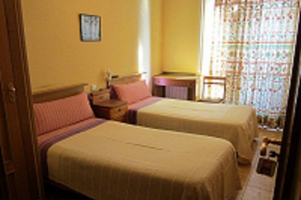 Habitación de hotel con dos camas, una mesita de noche de madera, una pequeña mesa y un ventanal con cortinas estampadas.
