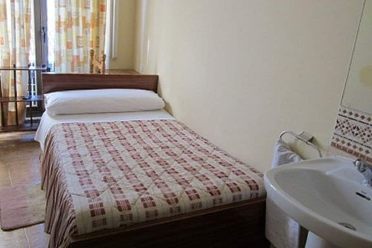 Un dormitorio con cama, una mesita de noche, una ventana con cortinas y un baño con lavamanos y espejo.